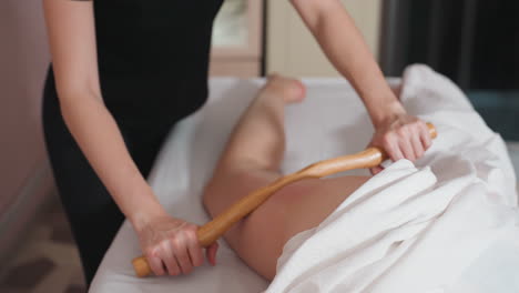 massage therapist using wooden stick to apply gentle pressure on client thigh during wellness session, performing smooth and steady movements in relaxing spa environment with soft linens