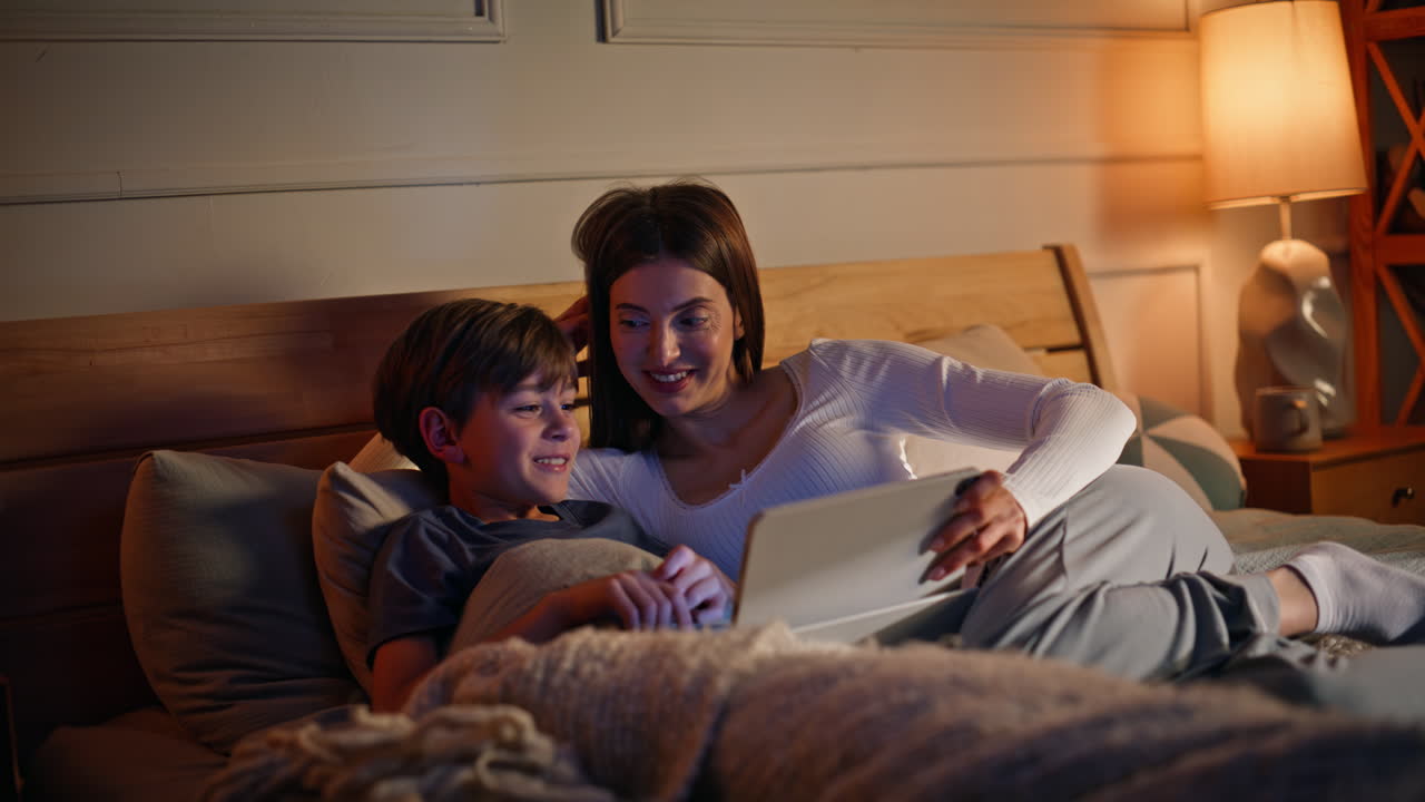 Woman holding tablet son lying bed dark apartment closeup. Smiling mother boy