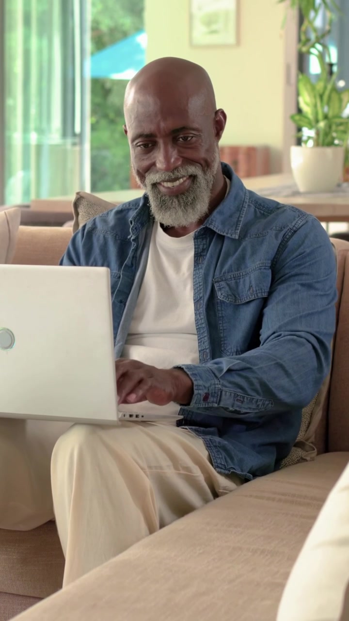 Vertical video: Typing man in denim shirt on sofa working on laptop while glass door drawing glance