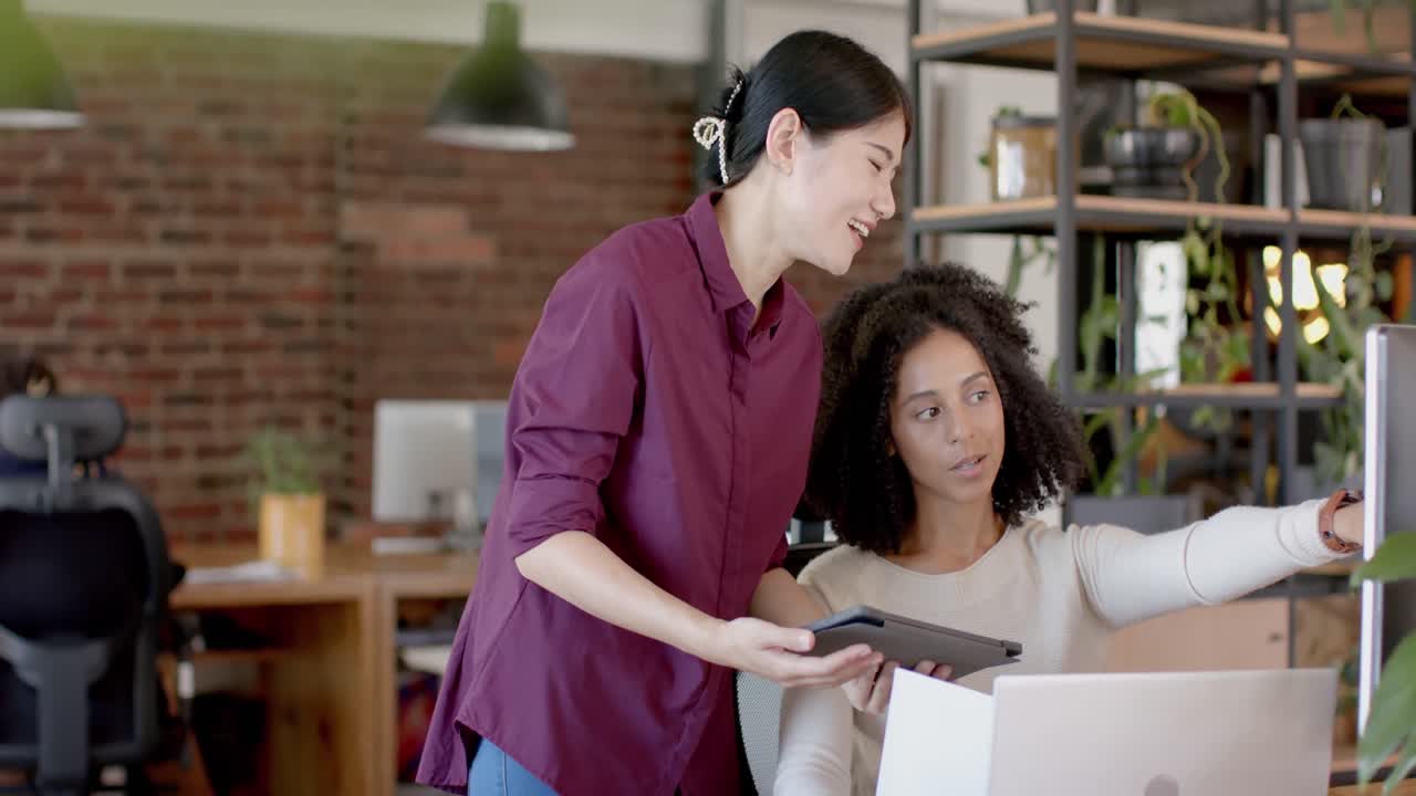 mujeres de negocios casuales felices y diversas usando tabletas, computadoras portátiles y hablando en la oficina en cámara lenta