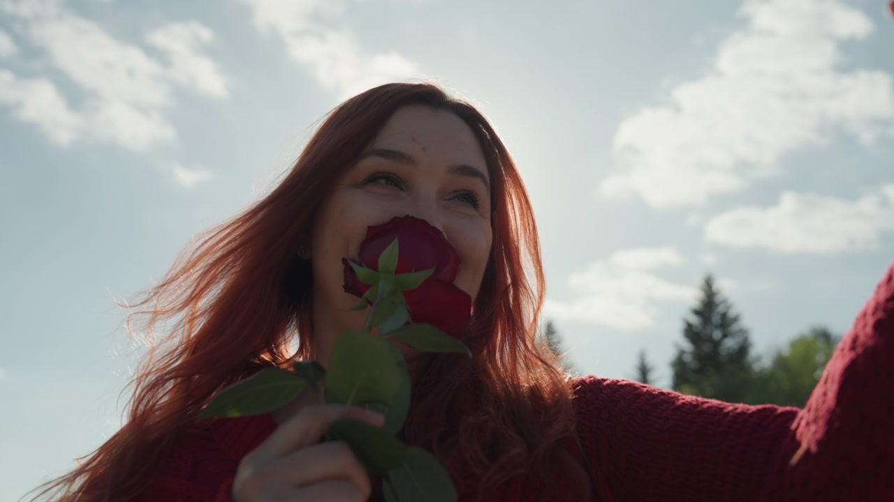 Kırmızı gül tutan ve akıllı telefonla dışarıda selfie çeken bir kadının yakın çekimi. Güneş ışığı, ağaçlar ve gökyüzü arka planıyla doğal bir ortamda uzun saçlarını vurgulayan bir lens parlaması oluşturuyor.
