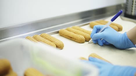 Unrecognizable-female-confectioner-hands-in-blue-gloves-write-some-marks-down-on-baking-parchment-under-threes-of-eclairs-or-profiteroles-layed-on-a-tray