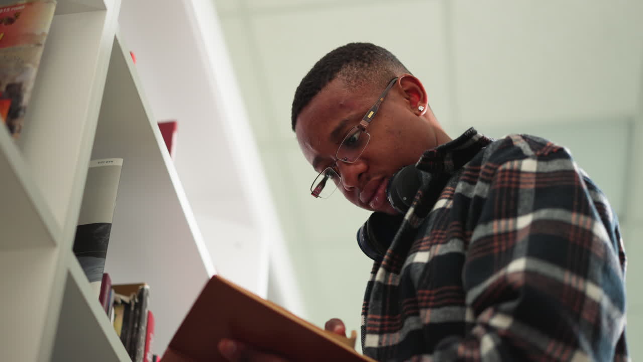 Man reads book in public library. African American reader with open textbook stands on ladder by bookcase. Serious student chooses scientific literature