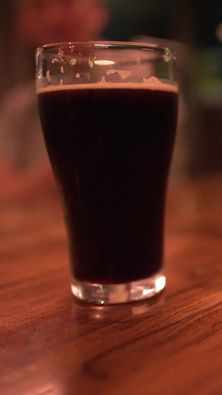 Ahşap bir pub masasında duran koyu renkli bir stout bira bardağının sabit yakın çekim görüntüsü.