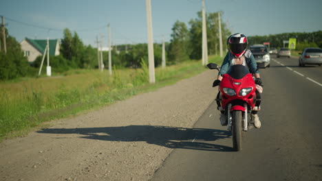 a female biker rides slowly on a paved road with cars moving in opposite directions, electric poles, trees, and distant buildings are visible in the background