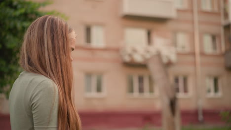 mujer joven con cabello largo y natural mirando hacia abajo pensativo mientras camina más allá de un edificio, ella parece contemplativa y cansada, la escena en un entorno urbano al aire libre con vegetación