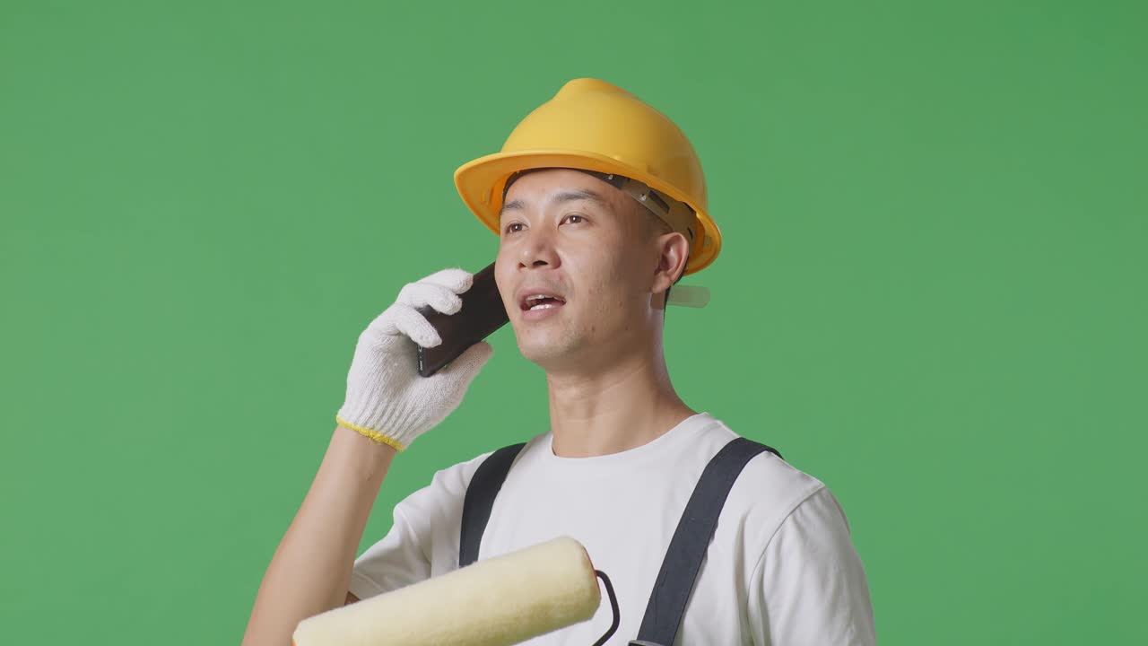 Close Up Of Side View Asian Man Painter Wearing Safety Helmet Talking On Smartphone And Looking Around While Standing In The Green Screen Background Studio