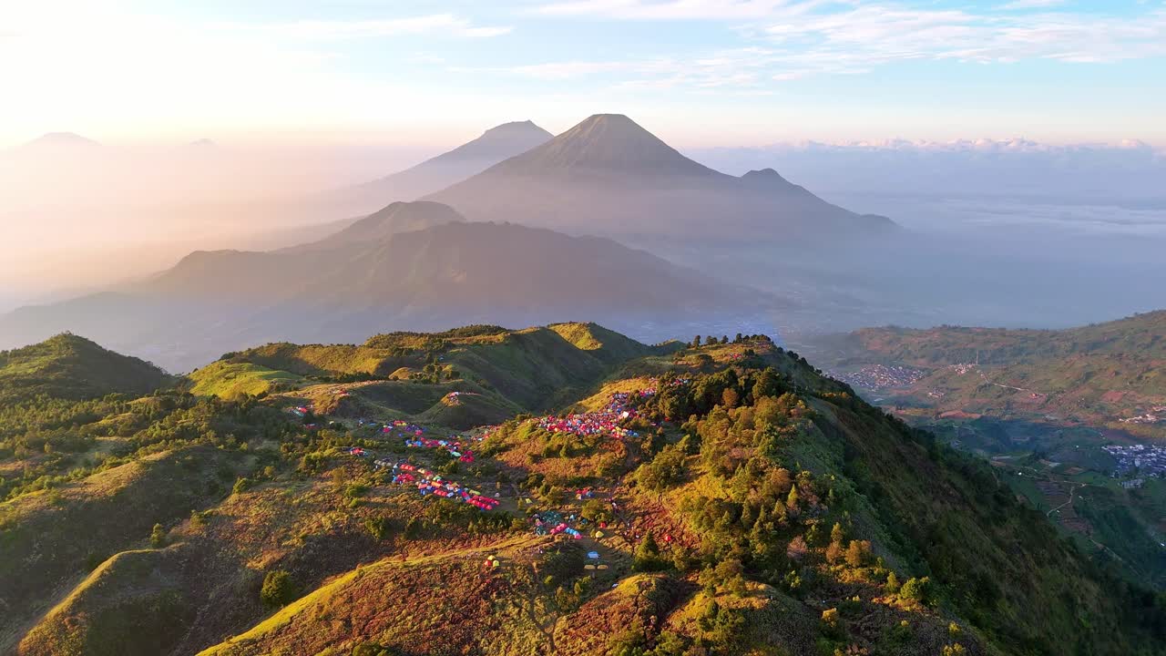 Ripresa cinematografica aerea dell'alba su una cima piena di campeggiatori. La luce soffusa del mattino bagna le colline e le tende, con sagome di montagne suggestive in lontananza. Monte Prau, Indonesia