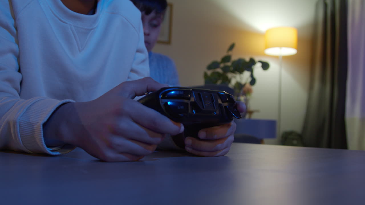 Close Up On Hands Of Two Young Boys At Home Playing With Computer Games Console On TV Holding Controllers Late At Night