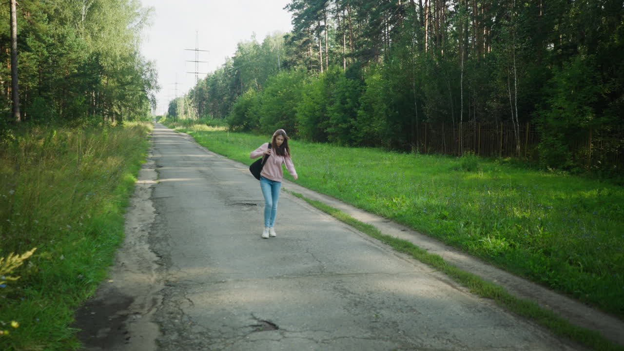 Young female freelancer in hoodie and jeans walking thoughtfully along empty cracked tarred road lined with green grass and trees, carrying bag over shoulder under daylight in quiet outdoor setting