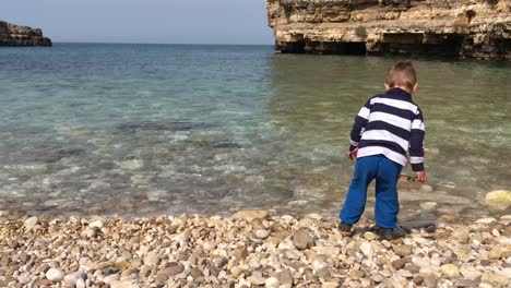 Niño-Pequeño-En-La-Playa-Arrojando-Rocas-Al-Agua-De-Mar,-Vacaciones-Infantiles-En-La-Playa