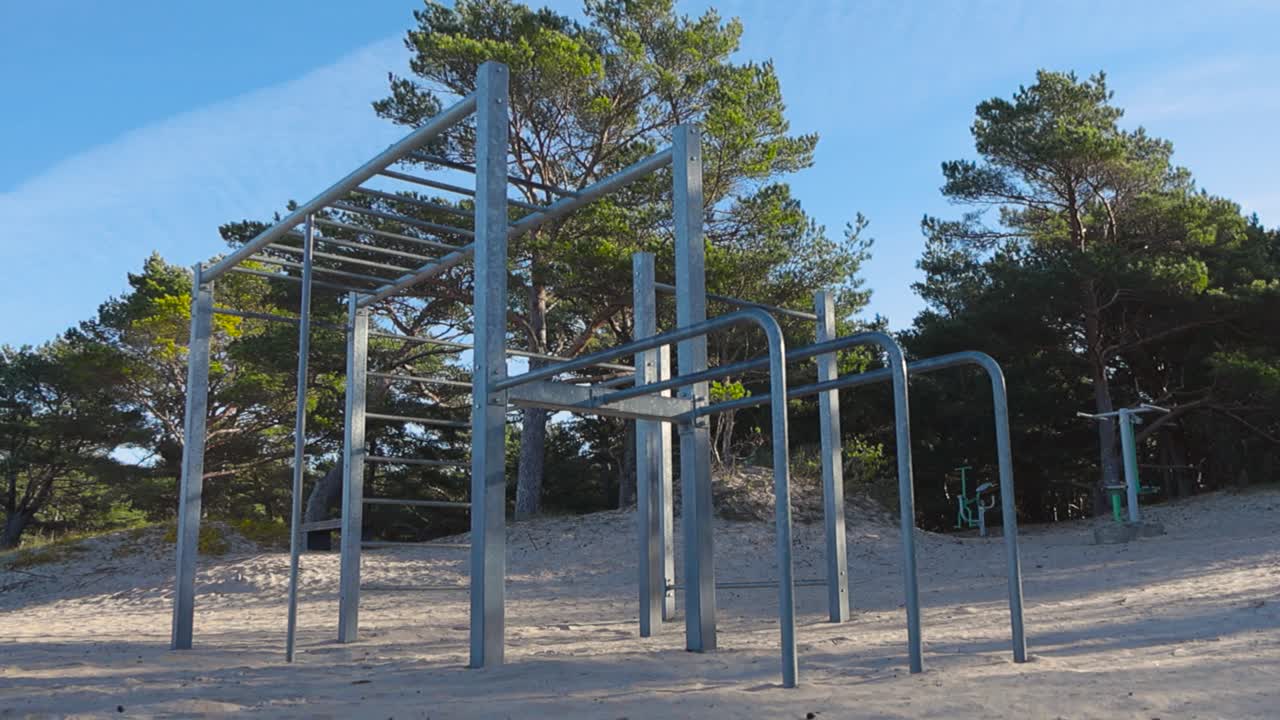 Outdoor gym workout place in a sandy beach during sunny day in summer or autumn in Vääna Jõesuu beach. Sky is blue and clear and tall pine trees are in the background that have sunlight on them.