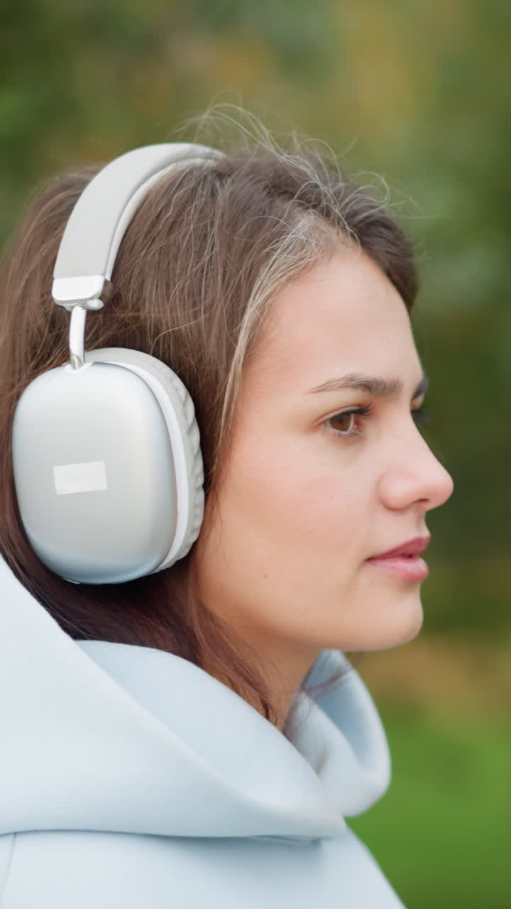 Close-up view of pretty lady in headphones listening to music in garden with blurred trees background, perfect for scenes depicting relaxation, outdoor activities, or enjoying nature with music