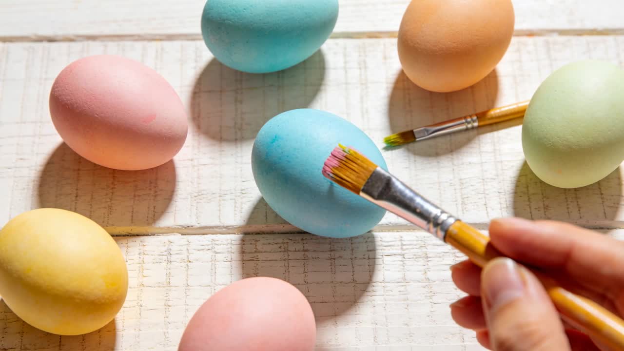 Una mano con pincel aplicando un toque de color a un huevo azul pastel sobre listones de madera, añadiendo una mancha rosa, con huevos cercanos.
