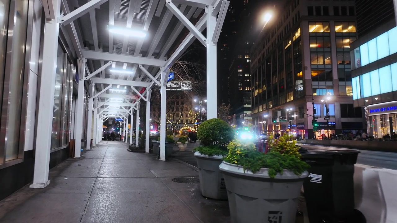 New York, USA, 25 dicembre 2025: Marciapiede coperto con fioriere a Midtown Manhattan di notte. Impalcatura bianca incornicia un corridoio di pavimentazione bagnata accanto a negozi, edifici per uffici e lampioni.