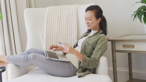 happy asian woman using laptop and holding credit card in bedroom, in slow motion