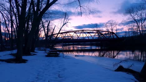 caminar a lo largo de la orilla de un río en un parque público verde al atardecer durante el invierno con nieve en el suelo, bancos y mesa de picnic, y un pequeño puente de acero arqueado rojo
