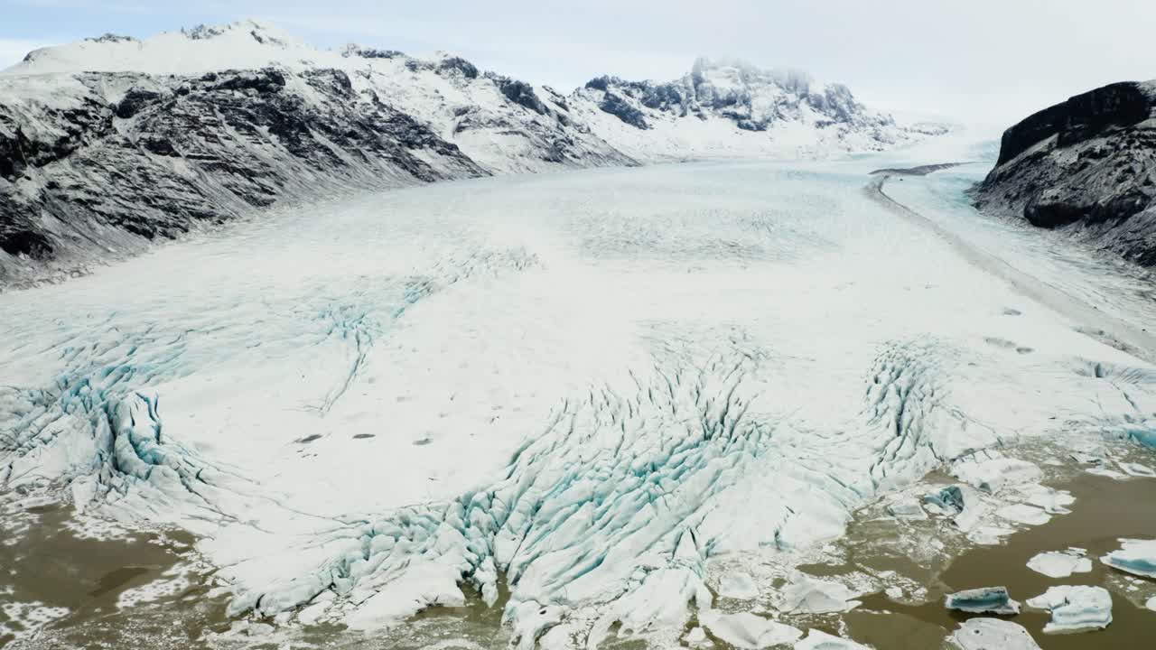 Aerial footage showcasing the icy expanse of Svinafellsjokull glacier transitioning into a serene muddy lagoon amidst dramatic mountain scenery.