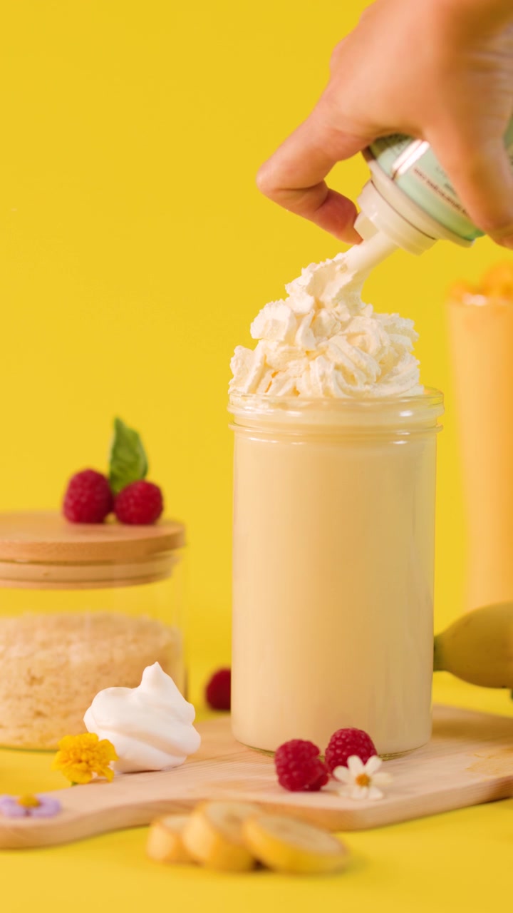 Una mano guarnisce un cremoso frullato di banana con panna montata in un allestimento alimentare da studio di colore giallo brillante