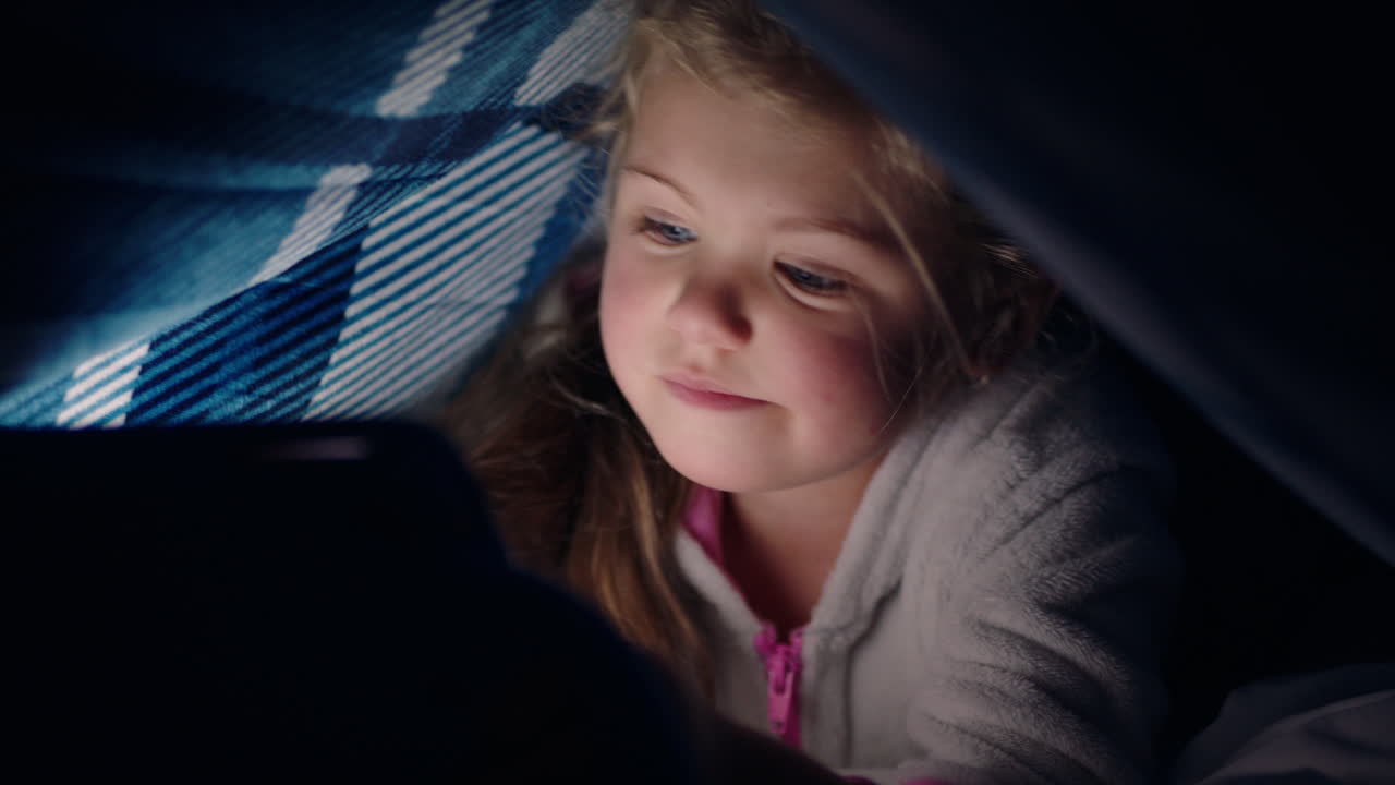 happy little girl using digital tablet computer under blanket enjoying learning on touchscreen technology playing games having fun at bedtime