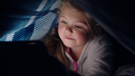 happy little girl using digital tablet computer under blanket enjoying learning on touchscreen technology playing games having fun at bedtime