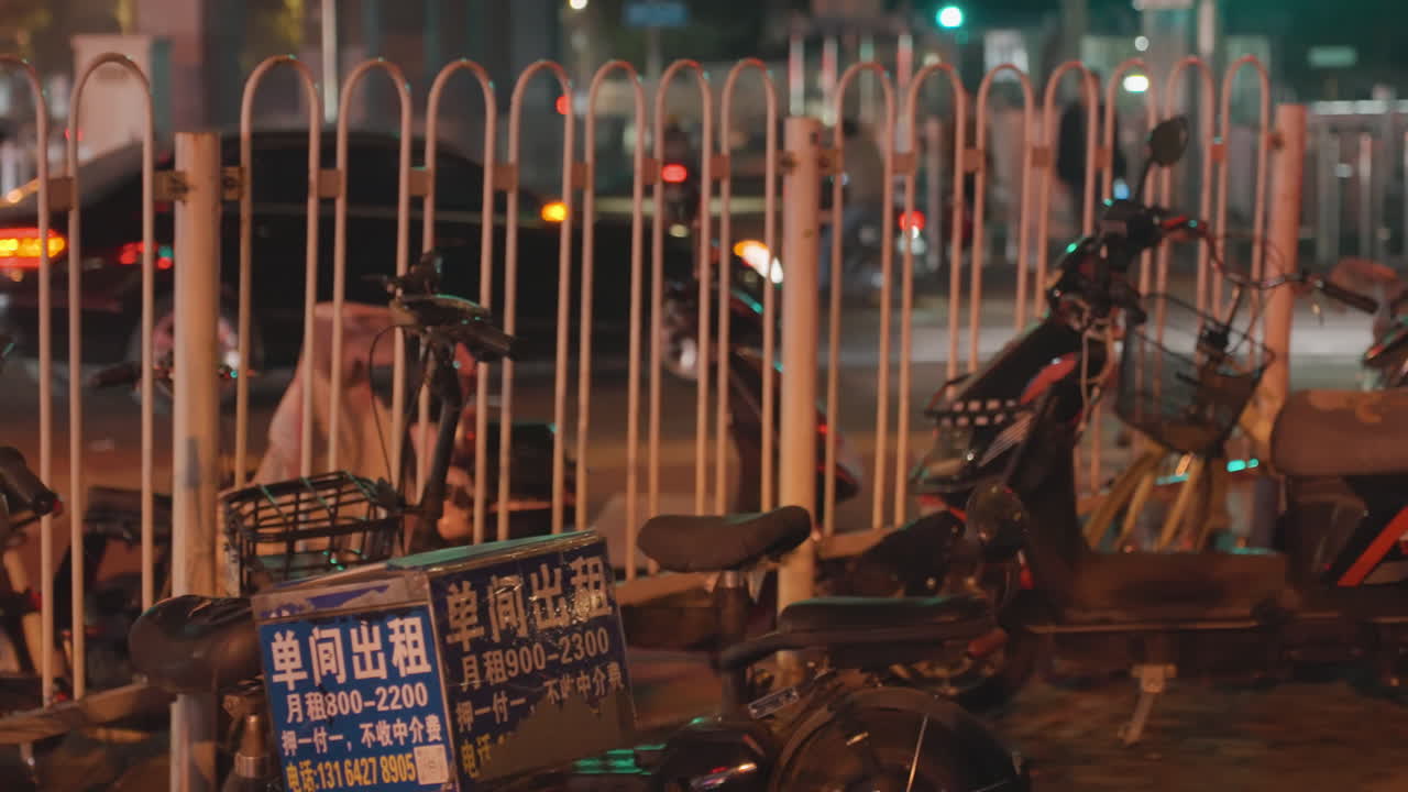 フェンス沿いに電動自転車が並ぶ夜の通り。暖かい街灯の光、配達用の箱、絡まった鍵と使い古されたシートが目立つ。遠くから聞こえる車のざわめき、反射する舗装路、控えめなネオンが、ゆっくりとした都市の鼓動を感じさせる。