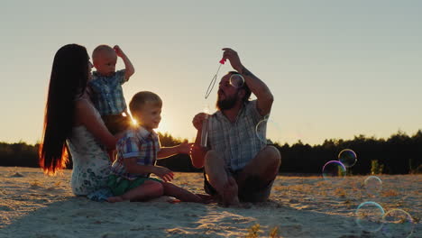 young parents and two sons of the family playing with soap bubbles at sunset