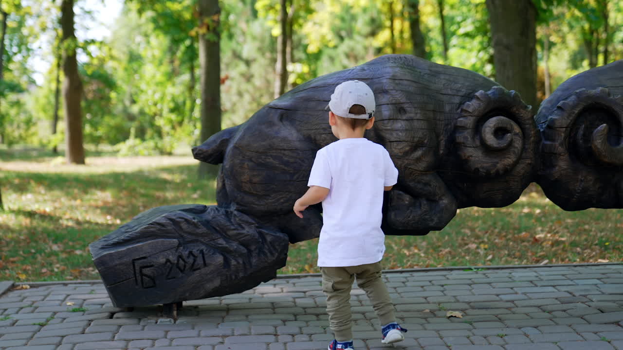 Active Caucasian toddler boy running around the wooden sculptures in the park. Kid touches the figures of bulls and taps by them.