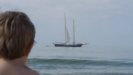 niño caucásico mirando un barco vintage navegando en la superficie del mar desde la distancia