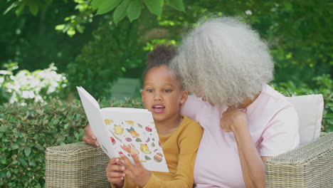 Grandmother-Sitting-Outdoors-In-Garden-With-Granddaughter-At-Home-Reading-Book-Together