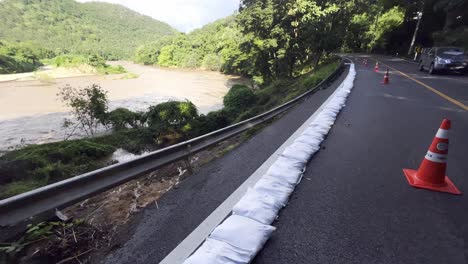 sandbags and red traffic cones lining the street along flowing river with high water level during flood season in thailand