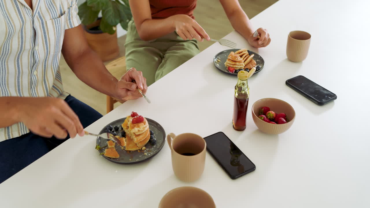 Coppia multietnica che fa colazione, mangiando pancake con caffè e frutti di bosco freschi a tavola, a casa