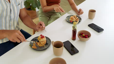 Enjoying breakfast, diverse couple eating pancakes with coffee and fresh berries at table, at home