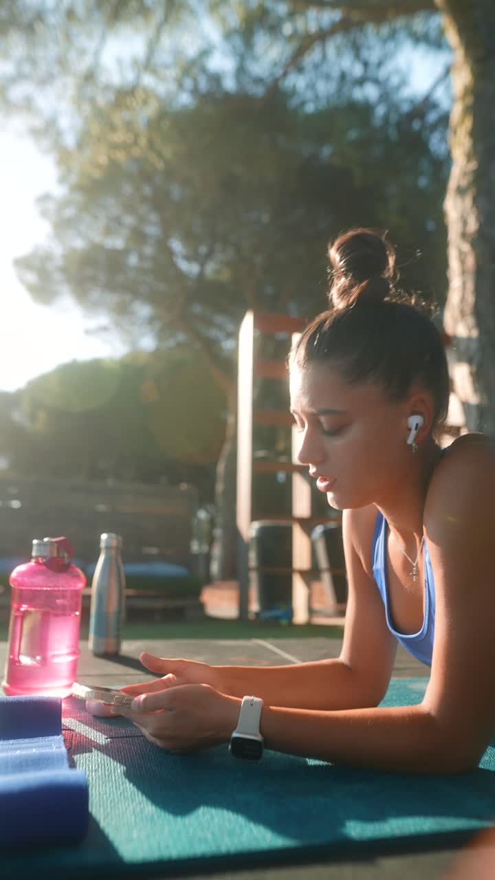 Woman resting on yoga mat outdoors with phone and water bottle