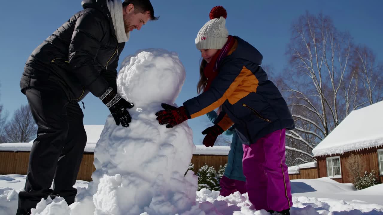 Low-angle video shot of a family building a snowman on a sunny winter day, capturing joyful