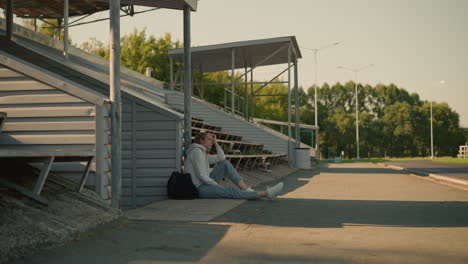 young woman seated alone on empty stadium bleachers, leaning against metal pole with thoughtful expression, sunlight filters through trees in background with shadows casted on the ground