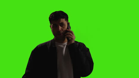 Young-Man-Talking-On-Mobile-Phone-Standing-Against-Green-Screen-Studio-Background-With-Low-Key-Lighting