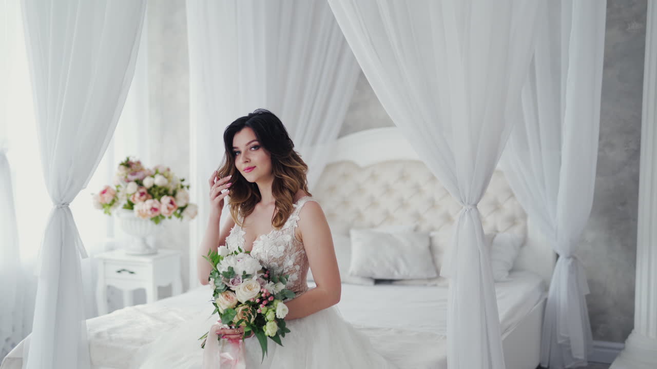 Beautiful young model in a white lace dress sits on a white bed. Gorgeous bride in wedding dress with flowers is sitting in the light bedroom.