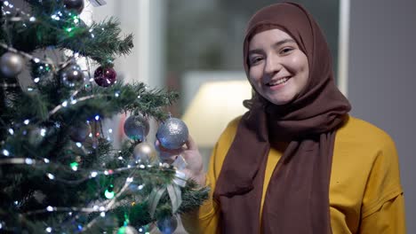 happy muslim young woman decorating christmas tree indoors turning to camera smiling. portrait of excited beautiful lady in hijab posing indoors on new year's eve. holidays season concept.