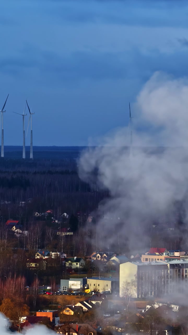 Smoke billows over a rural village with wind turbines in the background under a blue sky. Ideal for environmental themes or energy topics.