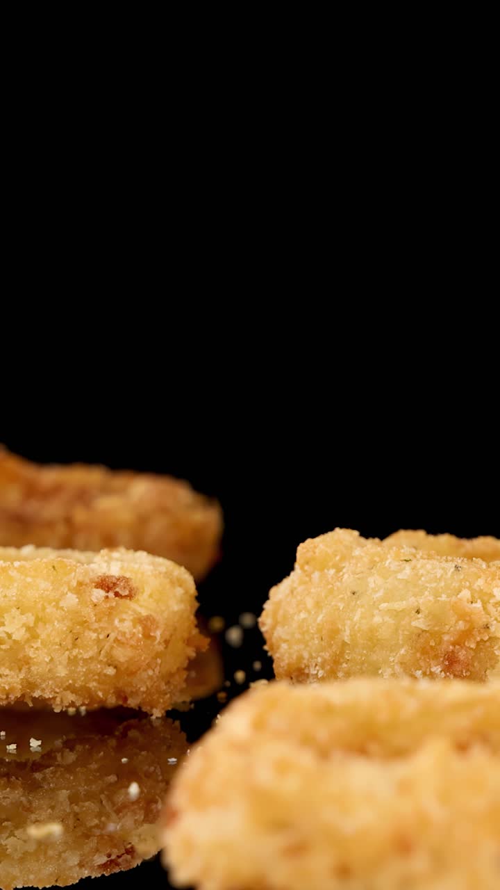 A golden, breaded onion ring falls onto a pile of onion rings against a black background, captured in slow motion with dramatic studio lighting