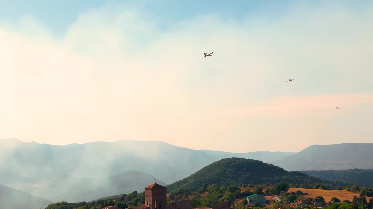 Luftbårne brandslukningsfly flyver over røgfyldte bakker og en landlig landsby under bekæmpelse af skovbrand.