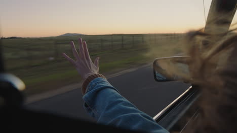 Junge-Frau-Im-Auto-Hält-Die-Hand-Aus-Dem-Fenster-Und-Spürt,-Wie-Der-Wind-Durch-Die-Finger-Weht.-Sie-Fährt-Auf-Einem-Roadtrip-Durch-Die-Landschaft-Und-Genießt-Die-Freiheit-Auf-Der-Straße-Bei-Sonnenuntergang