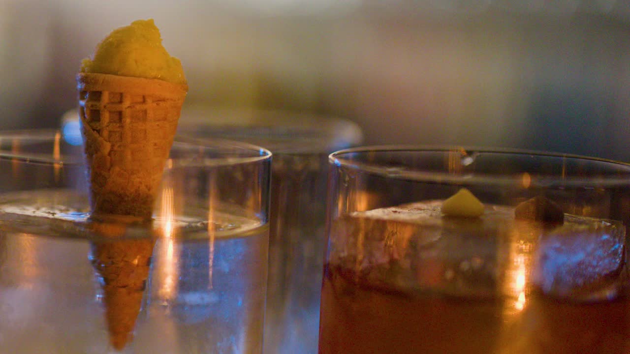 Close-up of cocktail glass with ice, mini cone garnish, warm bokeh lighting, shallow focus