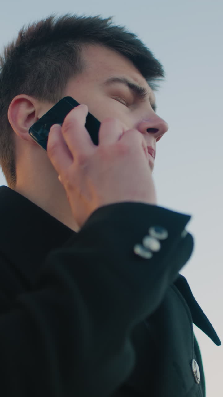 검은 코트와  셔츠를 입은 전문적인 남자가 은 도시 스카이라인을 배경으로 밖에서 드폰을 쳐다보고 집중과 결단력을 반영하고 있습니다.