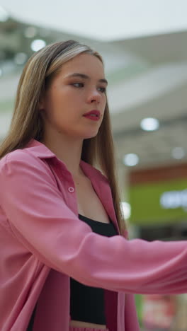 dama de vestido rosa con bolso de mano negro intensamente enfocada en el uso de una pantalla en un centro comercial bien iluminado, el fondo presenta luces borrosas y escaleras mecánicas, parece profundamente concentrada