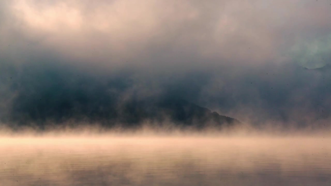Damp og skyer som beveger seg over en innsjø ved soloppgang, med en fjelltopp som stikker opp i det fjerne