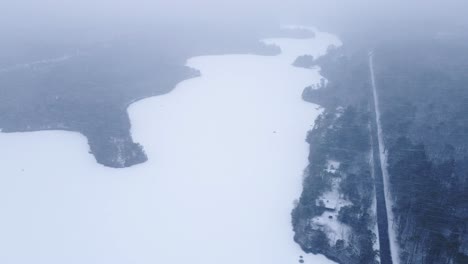 heavy snowstorm above frozen lake with aerial view of pine trees and asphalt road in winter