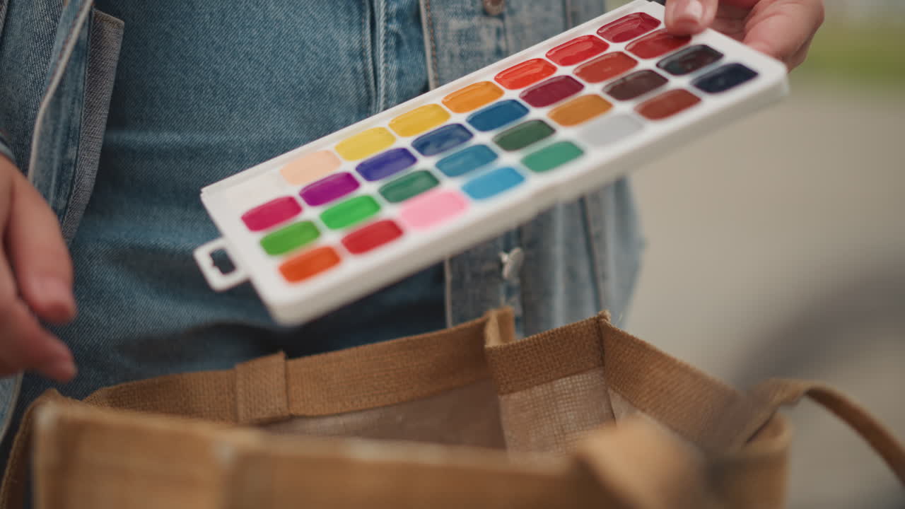 Mulher de jeans alcança uma bolsa de juta para pegar paleta de aquarela e pincel, close-up da mão e roupa casual de jeans, luz natural suave no banco, texturas táteis da sacola e do jeans, calma criativa