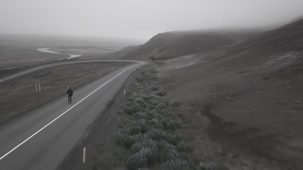 아이슬란드의 안개가 자욱하고 극적인 풍경 속에서 구불구불한 도로를 타고 내려가는 스케이터의 드론 샷